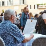 Błędy w nazwach dań po włosku, które zdradzają turystę w 10 sekund Starszy mężczyzna w kwiecistej koszuli czyta menu w rzymskiej kawiarni